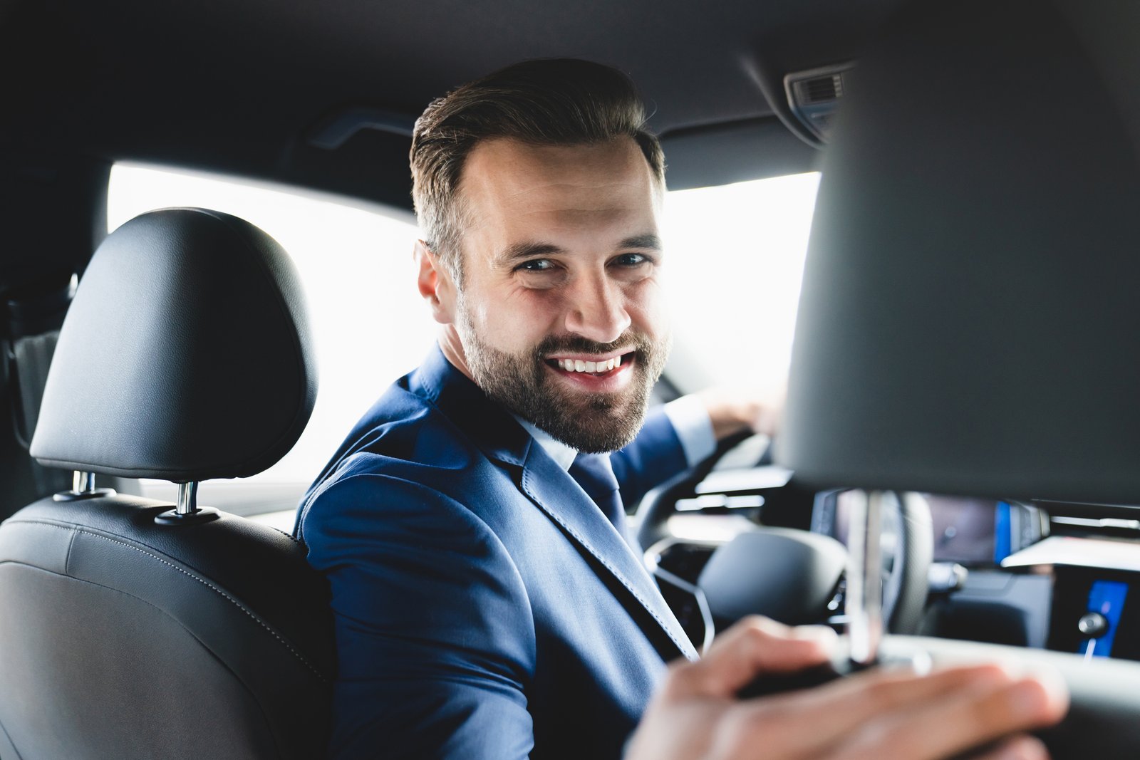 Taxi Driver Smiling While Turning Over To The Backseat Looking Talking With Clients. Successful Businessman Husband Looking At The Backseat At Dealer Shop Before Buying A Car Taxi Driver Smiling While Turning Over To The Backseat Looking Talking With Clients. Successful Businessman Husband Looking At The Backseat At Dealer Shop Before Buying A Car